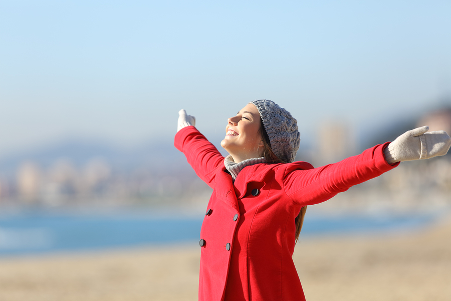 Happy woman wearing a red jacket breathing fresh air and raising arms on the beach in a sunny day of winter