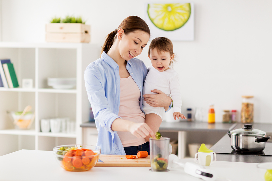 family, food, healthy eating, cooking and people concept - happy mother adding chopped vegetables to blender cup and holding little baby girl at home kitchen