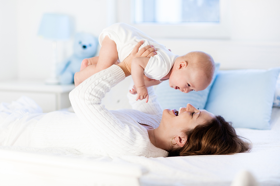 Mother and child on a white bed. Mom and baby boy in diaper playing in sunny bedroom. Parent and little kid relaxing at home. Family having fun together. Bedding and textile for infant nursery.